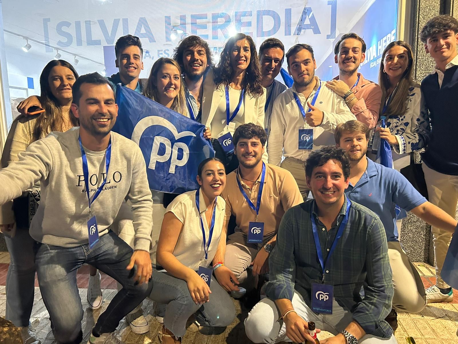 Grupo de jóvenes de NNGG Écija sonriendo juntos de noche, con una bandera del Partido Popular